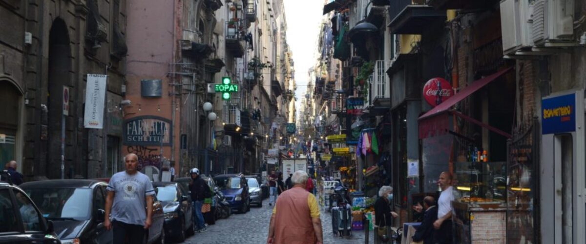 flaneur-porosidad-napoles Calle urbana de Nápoles con tránsito peatonal y actividades diversas, ilustración de la porosidad urbana y el flujo de la vida cotidiana en la ciudad.